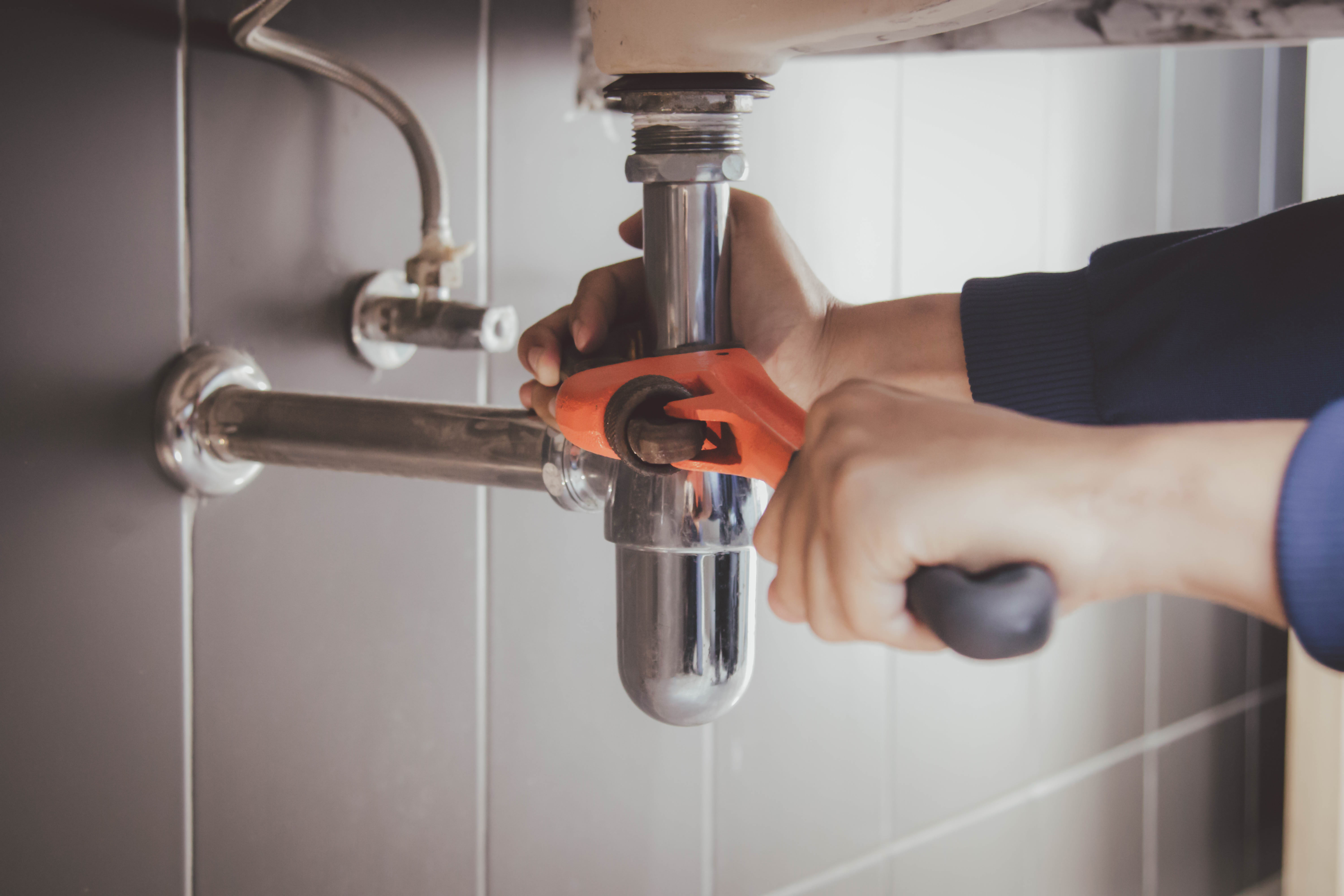 Plumber in orange gloves tightening chrome P-trap under sink with wrench